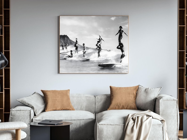 May include: Black and white photograph of a group of people surfing on a beach. The people are standing on surfboards and are in the process of riding waves. The photo is taken from a distance and shows the people in the water with the beach in the background.