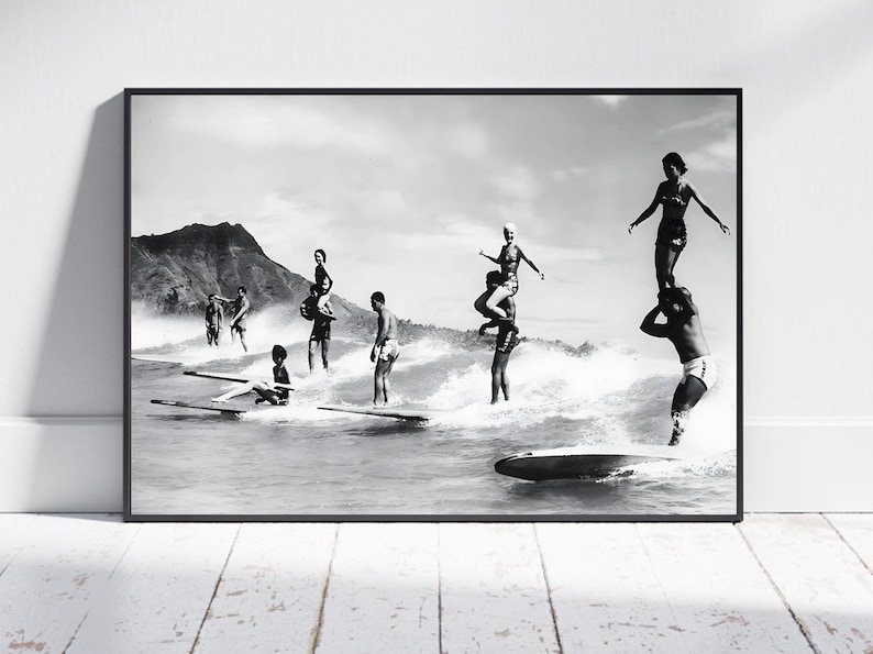 May include: Black and white photograph of a group of people surfing on a beach. The people are standing on surfboards and riding waves. There is a mountain in the background.
