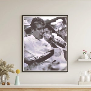 May include: A black and white photograph of a man in a chef's uniform drinking from a bottle while holding a frying pan.