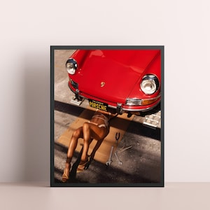 May include: A red vintage Porsche car with a California license plate that reads "PORSCHE" is lifted up on a jack. A person is lying underneath the car on a cardboard box.