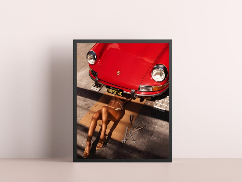 May include: A red vintage Porsche car with a California license plate that reads "PORSCHE" is lifted up on a jack. A person is lying underneath the car on a cardboard box.