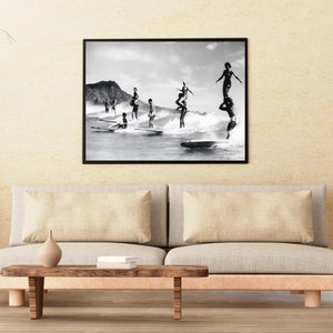 May include: Black and white photograph of a group of people surfing on waves. The photo is framed in a black frame and is hanging on a wall.