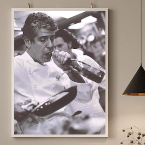 May include: A black and white photograph of a chef in a white chef's jacket pouring wine from a bottle into a pan. The chef is wearing a necklace and has a serious expression on their face.