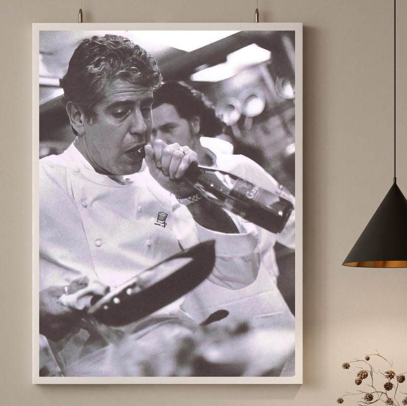 May include: A black and white photograph of a chef in a white chef's jacket pouring wine from a bottle into a pan. The chef is wearing a necklace and has a serious expression on their face.