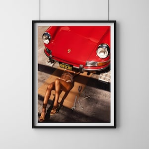 May include: A red vintage Porsche car is lifted on a jack, with a person lying underneath it. The car has a California license plate that reads "PORSCHE".