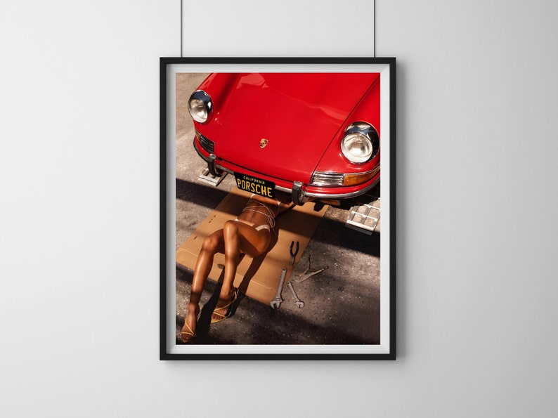 May include: A red vintage Porsche car is lifted on a jack, with a person lying underneath it. The car has a California license plate that reads "PORSCHE".