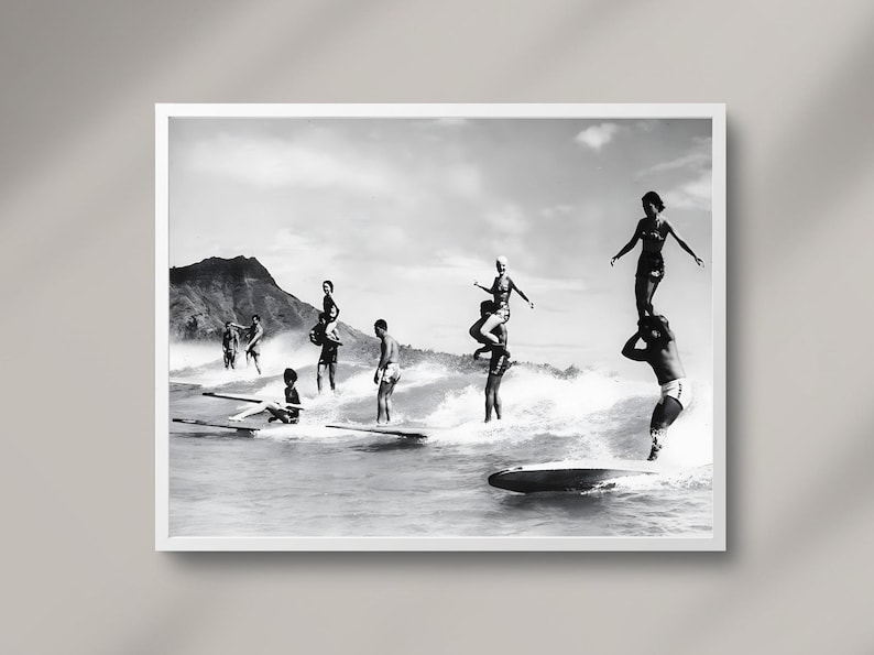 May include: A black and white photograph of a group of people surfing on a beach. The people are standing on surfboards and are riding the waves. The photo is taken from a distance and shows the people in the water.