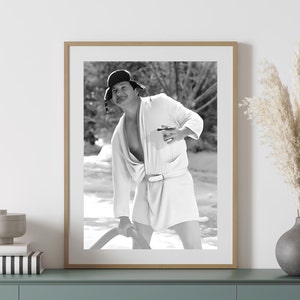 May include: Black and white photograph of a man wearing a white robe and a black fur hat. He is holding a drink in his right hand and smiling at the camera.