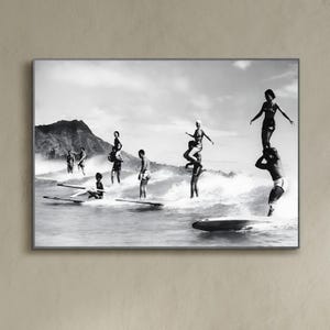 May include: A black and white photograph of a group of people surfing on a beach. The people are standing on surfboards and are riding waves. There is a mountain in the background.