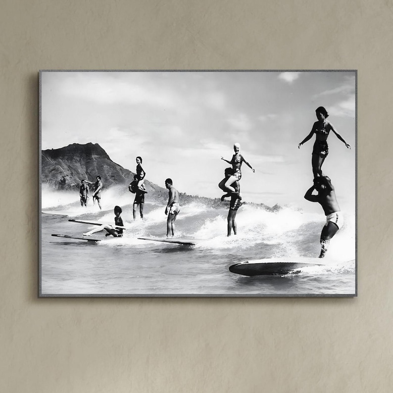 May include: A black and white photograph of a group of people surfing on a beach. The people are standing on surfboards and are riding waves. There is a mountain in the background.