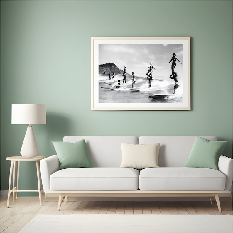 May include: Black and white photograph of a group of people surfing on a beach. The people are standing on surfboards and are in various poses. The beach is in the background and there are mountains in the distance.