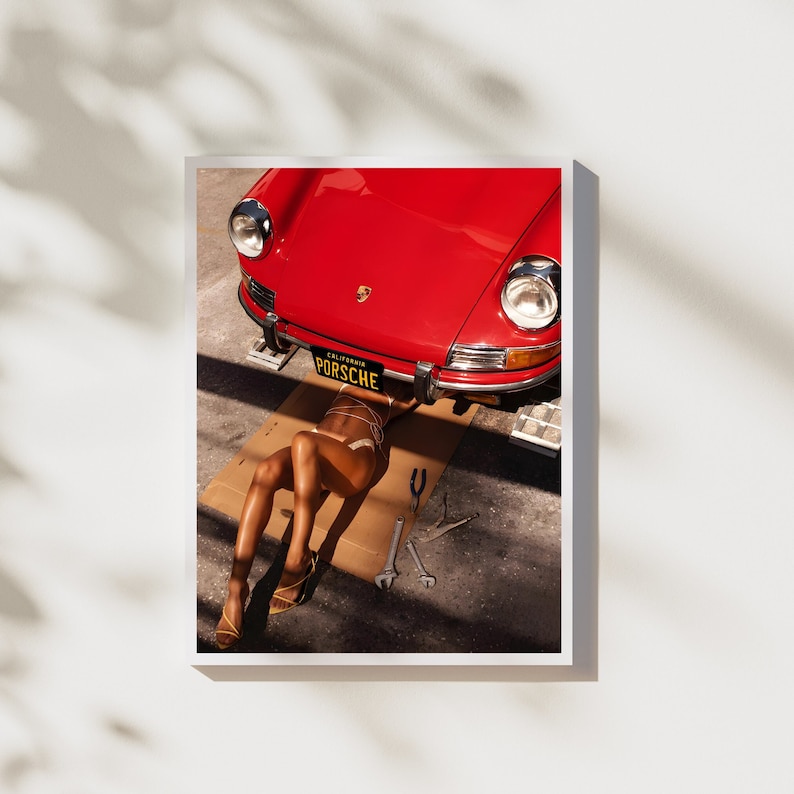 May include: A red vintage Porsche car is lifted up on a jack, with a person lying underneath it. The car has a California license plate that reads "PORSCHE".