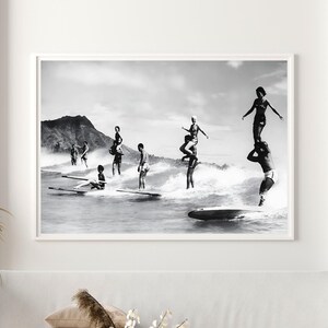 May include: Black and white photograph of a group of people surfing on a beach. The people are standing on surfboards and are riding the waves. There is a mountain in the background.