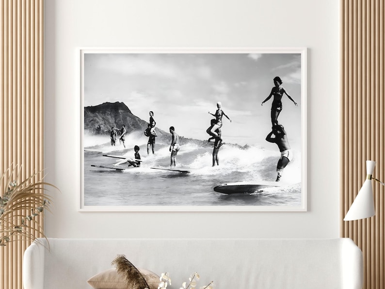 May include: Black and white photograph of a group of people surfing on a beach. The people are standing on surfboards and are riding the waves. There is a mountain in the background.