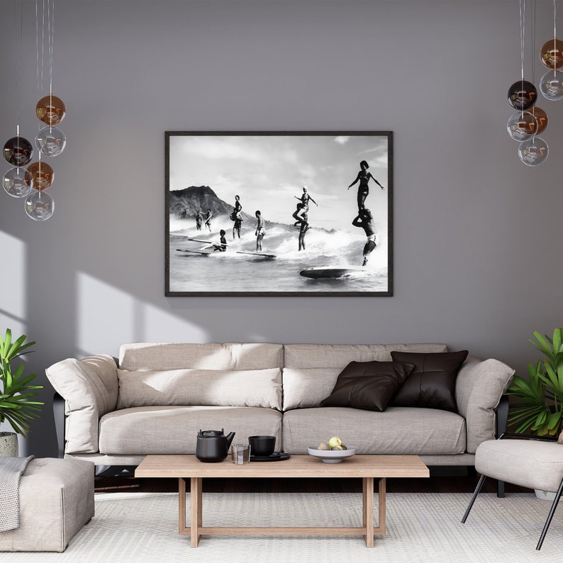 May include: Black and white photograph of a group of people surfing on a beach. The people are standing on surfboards and are in various poses. The photo is framed in a black frame.