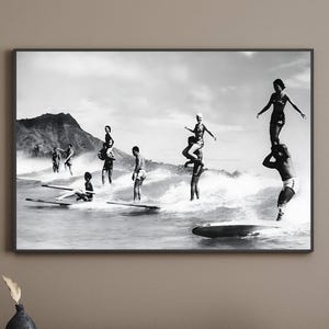 May include: Black and white photograph of a group of people surfing on a beach. The people are standing on surfboards and riding waves. There is a mountain in the background.