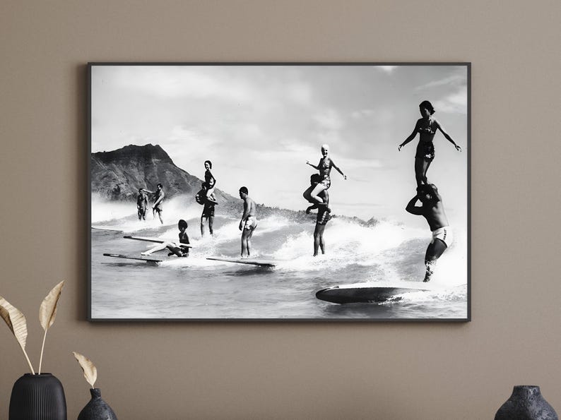 May include: Black and white photograph of a group of people surfing on a beach. The people are standing on surfboards and riding waves. There is a mountain in the background.