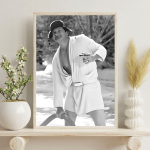 May include: A black and white photo of a person wearing a white robe and a black hat. The person is holding a cup in one hand and a hose in the other. The photo is taken outdoors.