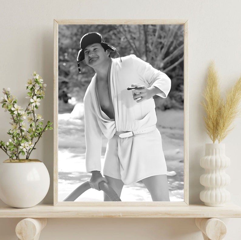 May include: A black and white photo of a person wearing a white robe and a black hat. The person is holding a cup in one hand and a hose in the other. The photo is taken outdoors.