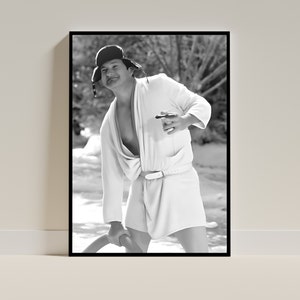 May include: A black and white photo of a person wearing a white robe and a black hat. The person is holding a cigar in one hand and a long object in the other hand. The person is standing in a snowy or icy environment.