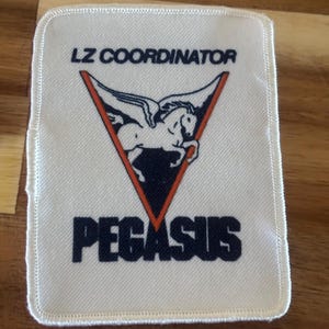 Peut inclure: Un écusson rectangulaire blanc avec le texte "LZ COORDINATOR" au-dessus d'un graphique de cheval ailé dans un triangle rouge et noir. Le mot "PEGASUS" est imprimé en dessous en bleu marine.
