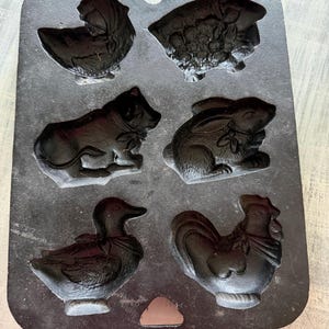 Peut inclure: Un moule de cuisson rectangulaire en fonte noire comportant six cavités en forme d'animaux. Le moule présente des empreintes d'un oiseau, d'un mouton, d'un chien, d'un lapin, d'un canard et d'un coq, chacun détaillé avec des textures et des nœuds.