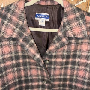 Könnte beinhalten: Eine rosa, grau und schwarz karierte Pendleton-Jacke. Die Jacke hat einen Reverskragen, drei Knöpfe und ein Etikett mit der Aufschrift "Pendleton". Die Jacke ist Größe 10.