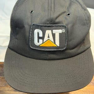 Peut inclure: Une casquette de baseball noire dotée d'un écusson rectangulaire sur le panneau avant avec le texte CAT en lettres blanches au-dessus d'un triangle jaune. La casquette présente une calotte structurée avec des œillets et une visière courbée.
