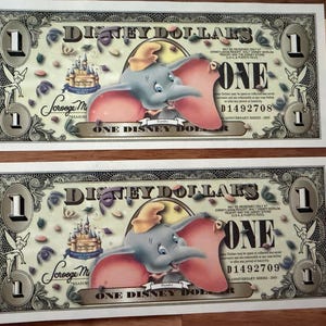 Puede incluir: Dos billetes de un dólar Disney Dollars con el elefante Dumbo en el centro, el texto Disney Dollars, el número 1 en las esquinas, una ilustración de un castillo, Campanilla y los números de serie D1492708 y D1492709 impresos en cada uno.