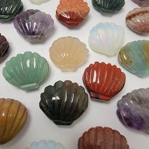 May include: A collection of carved gemstone seashell figurines in various colors, including green, purple, red, and white. The seashells are arranged on a white surface, showcasing their intricate details and natural variations.