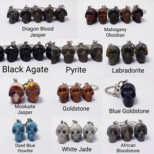 May include: A collection of 10 different skull-shaped keychains made from various types of stones, including Dragon Blood Jasper, Mahogany Obsidian, Black Agate, Pyrite, Labradorite, Mookaite Jasper, Goldstone, Blue Goldstone, Dyed Blue Howlite, and White Jade. Each keychain features a detailed skull carving and a silver key ring.