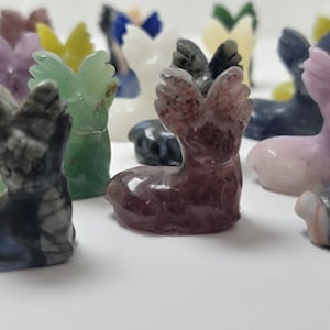 May include: A collection of small, carved deer figurines made from various types of polished stones. The deer have wings and are in a variety of colors, including purple, green, pink, and black.