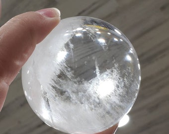 May include: A clear quartz crystal sphere with a cloudy, white interior. The sphere is being held in a hand.