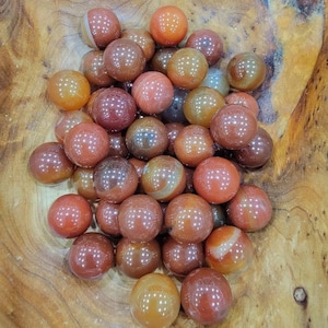 May include: A collection of carnelian tumbled stones in various shades of orange and red. The stones are smooth and polished, and they are arranged in a pile on a wooden surface.