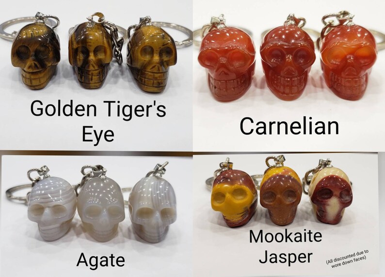May include: Three sets of three skull-shaped keychains made of different types of stone. The first set is golden tiger's eye, the second is carnelian, and the third is agate. The last set is mookaite jasper. All keychains are discounted due to worn down faces.