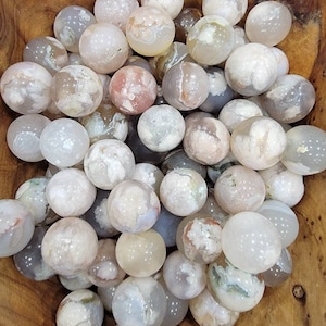 May include: A collection of white, gray, and brown agate spheres. The spheres are smooth and polished, with intricate patterns of white and gray swirls. The spheres are arranged in a bowl, and the background is a wooden surface.