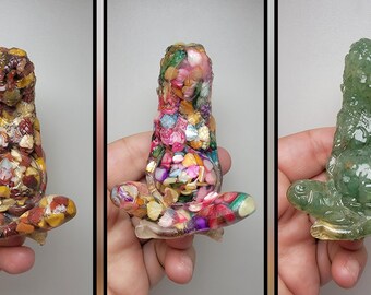 May include: Three resin sculptures of a pregnant woman in a seated meditation pose. The sculptures are made with embedded colorful stones and crystals. The first sculpture is a mix of red, orange, and yellow stones. The second sculpture is a mix of pink, purple, and green stones. The third sculpture is a solid green color.