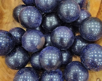 May include: A collection of polished blue sandstone spheres with sparkling inclusions. The spheres are of varying sizes and are arranged in a wooden bowl. The spheres have a deep blue color with reflective, glitter-like particles.
