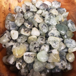 May include: A bowl filled with tumbled green aventurine stones. The stones are smooth and polished, with a translucent green color and black inclusions.