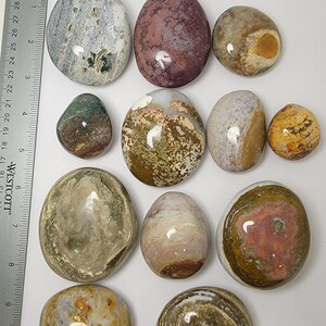 Puede incluir: Doce piedras lisas y pulidas con varios patrones y colores, incluyendo marr&oacute;n, blanco, gris, rosa y amarillo. Las piedras est&aacute;n dispuestas en una fila sobre una superficie blanca, con una regla en el fondo.