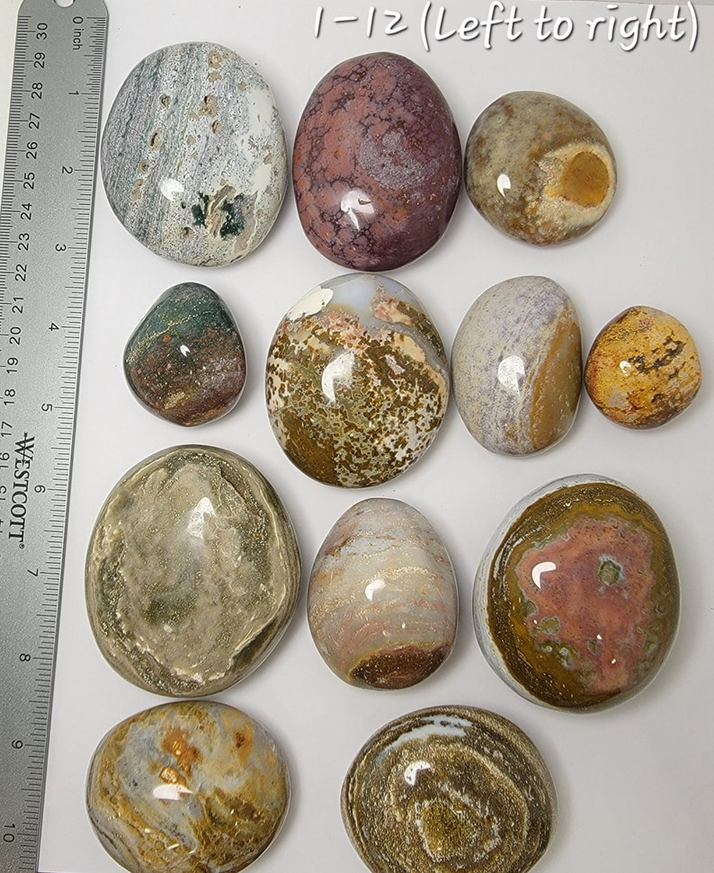 Puede incluir: Doce piedras lisas y pulidas con varios patrones y colores, incluyendo marr&oacute;n, blanco, gris, rosa y amarillo. Las piedras est&aacute;n dispuestas en una fila sobre una superficie blanca, con una regla en el fondo.