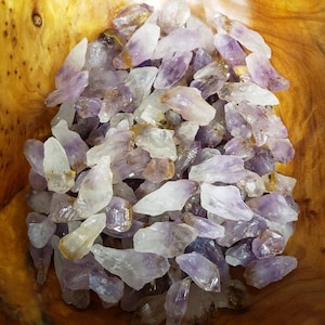May include: A bowl filled with amethyst crystals, some of which have a golden hue. The crystals are various sizes and shapes, with a pointed or jagged edge. The crystals are in a wooden bowl.