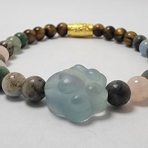 May include: A multi-colored beaded bracelet with a light blue paw charm. The bracelet features beads of tiger's eye, green aventurine, rose quartz, and aquamarine.