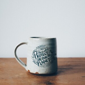 May include: White ceramic mug with a black and white illustration of flowers and the text "Protect Our Planet".