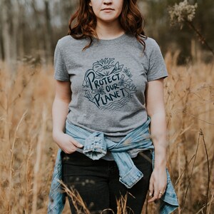 May include: A woman wearing a gray t-shirt with the text "Protect Our Planet" in a blue circle with a floral design. She is wearing a blue denim jacket tied around her waist and black jeans. She is standing in a field of tall, dry grass.