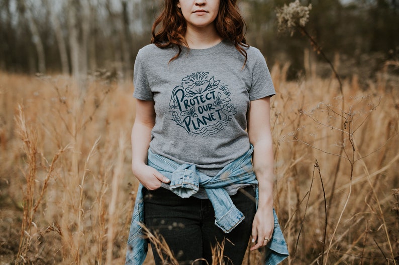 May include: A woman wearing a gray t-shirt with the text "Protect Our Planet" in a blue circle with a floral design. She is wearing a blue denim jacket tied around her waist and black jeans. She is standing in a field of tall, dry grass.