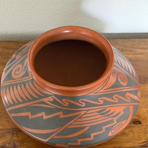 May include: A wide-mouthed terracotta pottery vessel featuring a rounded body and a flat rim. The exterior surface is decorated with intricate, hand-painted geometric patterns in matte black and natural clay orange, showcasing sharp zig-zag lines.