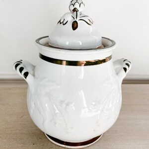 May include: White ceramic sugar bowl with a gold trim and a lid featuring a floral design. The bowl has two handles and a raised floral pattern.