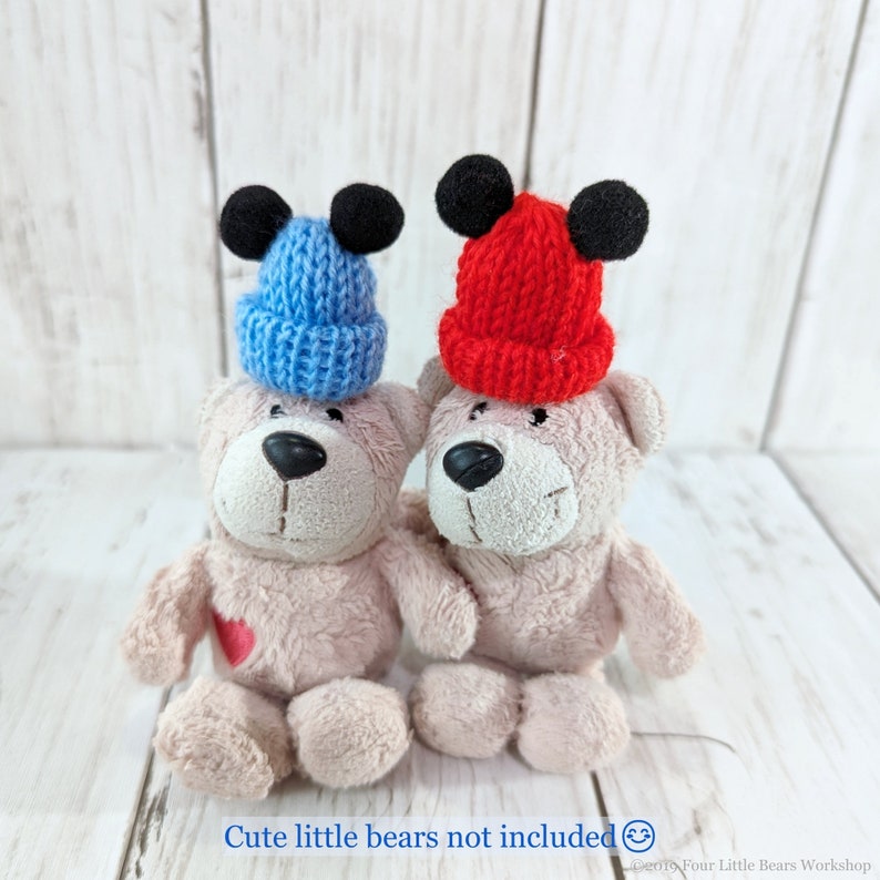 Pu&ograve; includere: Due cappelli di lana con le orecchie nere. Un cappello &egrave; rosso e l'altro &egrave; blu. I cappelli sono posizionati sopra due orsacchiotti.