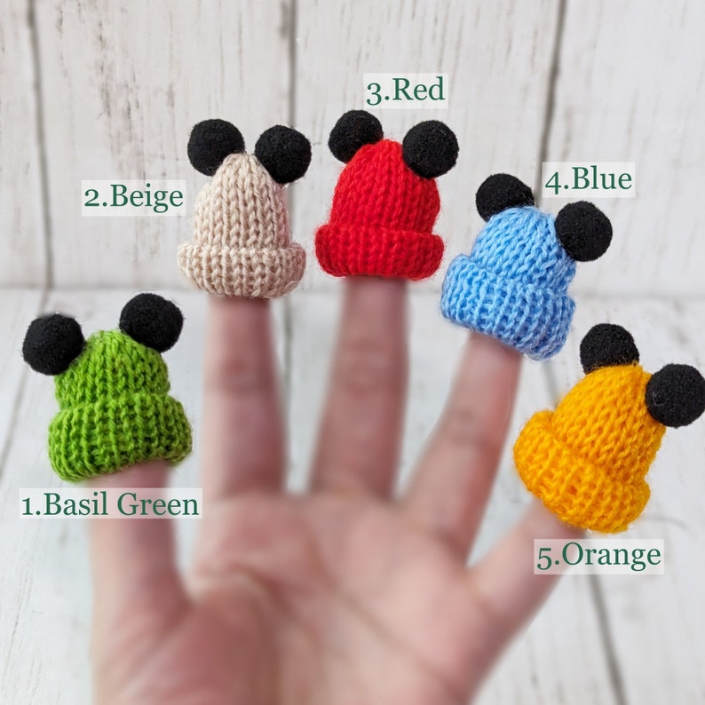 Pu&ograve; includere: Cinque pupazzi per dita a forma di berretti di lana con orecchie di Topolino. I berretti sono di colore verde basilico, beige, rosso, blu e arancione. Il testo "1. Basil Green", "2. Beige", "3. Red", "4. Blue" e "5. Orange" &egrave; scritto sotto ogni berretto.